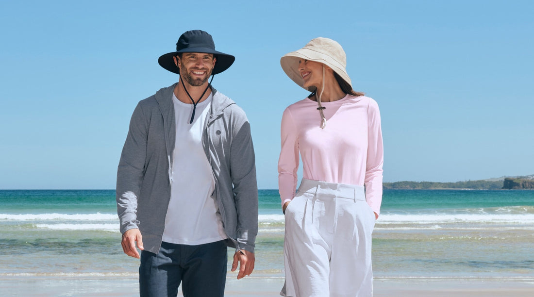 A man and a woman walking on a beach wearing Solbari sun huts and sun protective clothing.