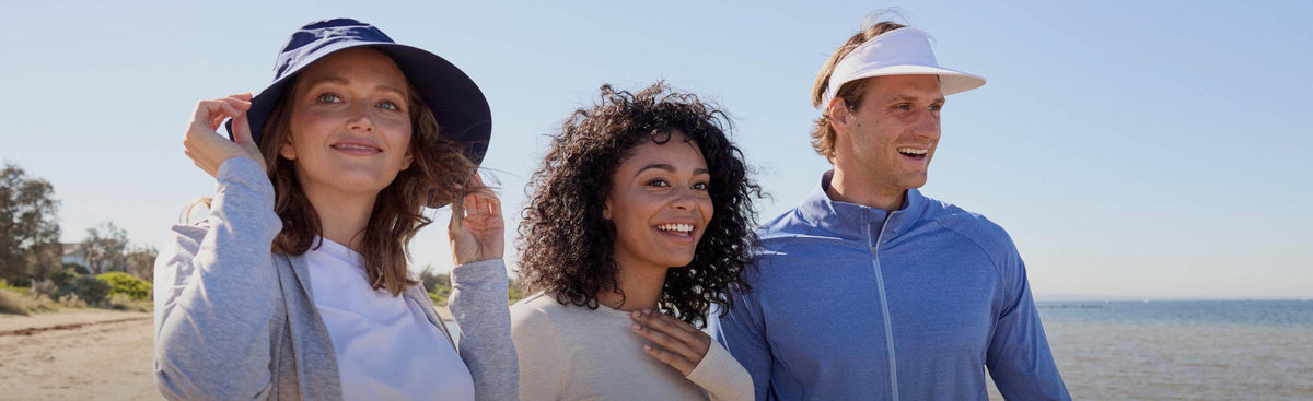 Three people standing on a beach wearing sun-protective clothing and hats.